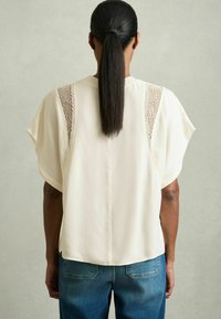 Cremfarbene Bluse mit kurzen Fledermausärmeln, SpitzenfDetail an den Seiten, weichem Stoff und geradem Rückendesign, kombiniert mit blauen Jeans.