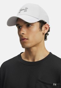 Balta cepure ar izliekto maliņu, uz kuras priekšpuses ir sudraba Under Armour logo. Valkāta ar teksturētu melnu krekli.