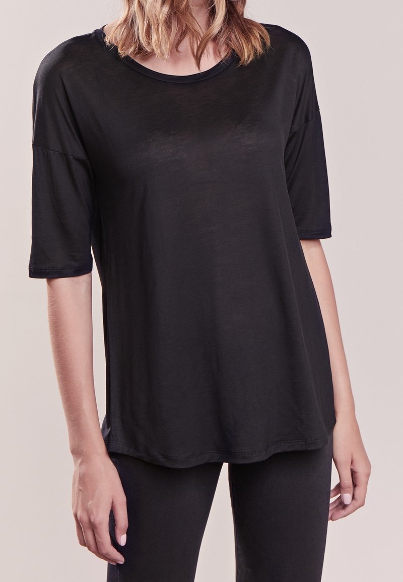 Femme portant un haut noir ample à manches courtes et un pantalon noir, debout devant un fond neutre.