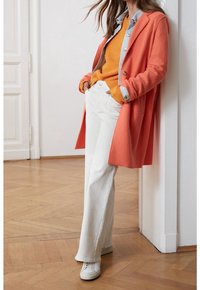 Cappotto di lana arancione, maglione arancione a maglia, camicia di jeans chiaro, pantaloni bianchi a gamba larga e sneakers bianche. Tessuto liscio e design a strati.