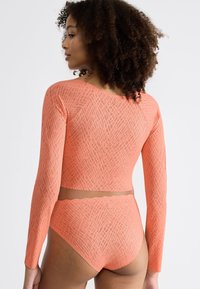 Kobieta z kręconymi włosami, ubrana w koralowy długa rękaw crop top oraz dopasowane high-waisted figi z delikatnym teksturowanym wzorem.
