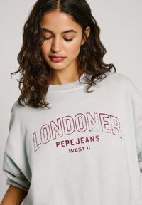 Šviesiai pilkas perteklinis džemperis su užrašu "LONDONER PEPE JEANS WEST II" tamsiai raudona spalva. Turi apvalią kaklo iškirptę ir laisvus rankogalius.