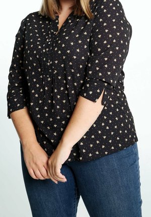 Osoba w czarnej bluzce z długim rękawem z drobnym kwiatowym wzorem, w połączeniu z niebieskimi dżinsami, ręce splecione z przodu.