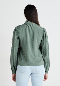 Blusa verde con un motivo a pois testurizzato, maniche lunghe a sbuffo e colletto rotondo, realizzata in tessuto leggero.