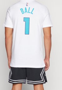 Camiseta de algodón blanca con "BALL" y el número "1" en un vibrante turquesa con una textura punteada, llevada con pantalones cortos de malla negros y acentos blancos.