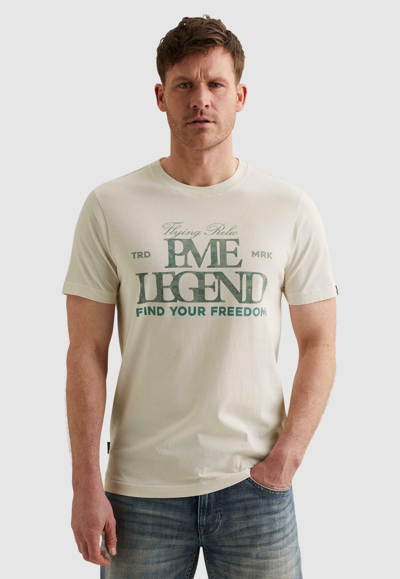 Homme portant un t-shirt beige clair avec le texte vert « Flying Relic PME Legend Find Your Freedom » et un jean bleu, debout avec une main dans la poche.