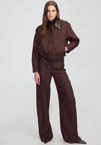 Giacca a quadri marrone con colletto a contrasto, abbinata a pantaloni a vita alta e a gamba larga, che mostrano un tessuto testurizzato e un motivo sottile.