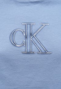 Lyseblå bomullsstoff med brodert "CK" i sølv. Jevn tekstur med et klassisk design, plassert sentralt på plagget.