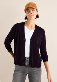 Dunkellila Strickjacke mit Knopfverschluss, gerippten Bündchen, getragen über einem weißen Hemd, kombiniert mit hellgrauen Jeans und einer braunen Mütze. Glatte Textur.
