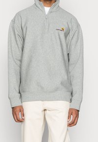 Sudadera de forro polar gris con cuello de media cremallera, puños acanalados y cinturilla. Presenta un pequeño logo en burdeos y amarillo en el lado izquierdo del pecho.