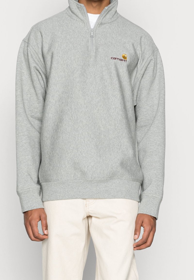 Sudadera de forro polar gris con cuello de media cremallera, puños acanalados y cinturilla. Presenta un pequeño logo en burdeos y amarillo en el lado izquierdo del pecho.