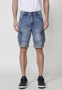 Denim cargo shorts, lichtblauw met een vervaagde textuur, met zijzakken, opgerolde zomen en een knoopsluiting. Gecombineerd met zwarte sneakers.