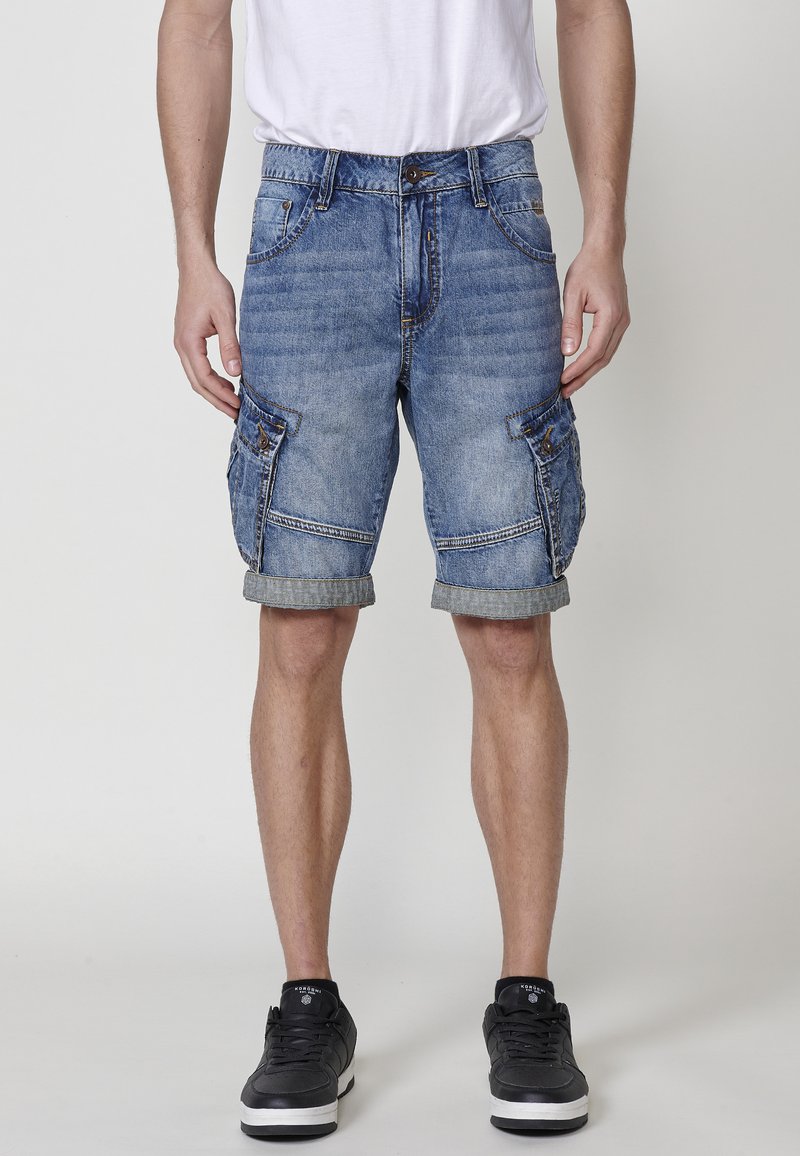 Denim cargo shorts, lichtblauw met een vervaagde textuur, met zijzakken, opgerolde zomen en een knoopsluiting. Gecombineerd met zwarte sneakers.