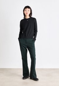Pull en maille noire avec un col rond, associé à un pantalon droite vert foncé. Le modèle se tient avec les mains dans les poches, portant des chaussures noires.