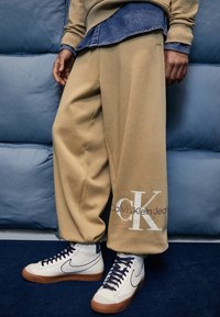 Beige oversized sweaterbroek met het logo "Calvin Klein Jeans", gecombineerd met witte high-top Nike sneakers. Zachte stof, ontspannen pasvorm.