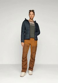 Pufi sötétkék dzseki, mintás zöld pulóver és barna cargo nadrág. Az öltözet funkcionális kialakítású, több zsebbel rendelkezik, bézs cipővel viselve.