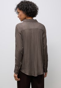 Chemise marron à manches longues avec un col, en tissu texturé, fentes latérales et poignets boutonnés. Le détail du pli au dos améliore le mouvement et l'ajustement.