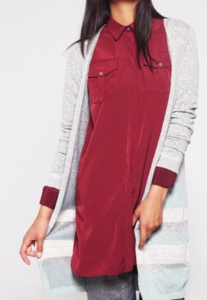 Kobieta nosząca długi szary kardigan w paski na czerwonej sukience koszulowej z dwoma kieszeniami na piersiach, stojąca na białym tle.