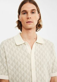Korte mouwen polo shirt in crème met een textuurpatroon van bladeren, voorzien van een puntige kraag en een knoopsluiting aan de voorkant.