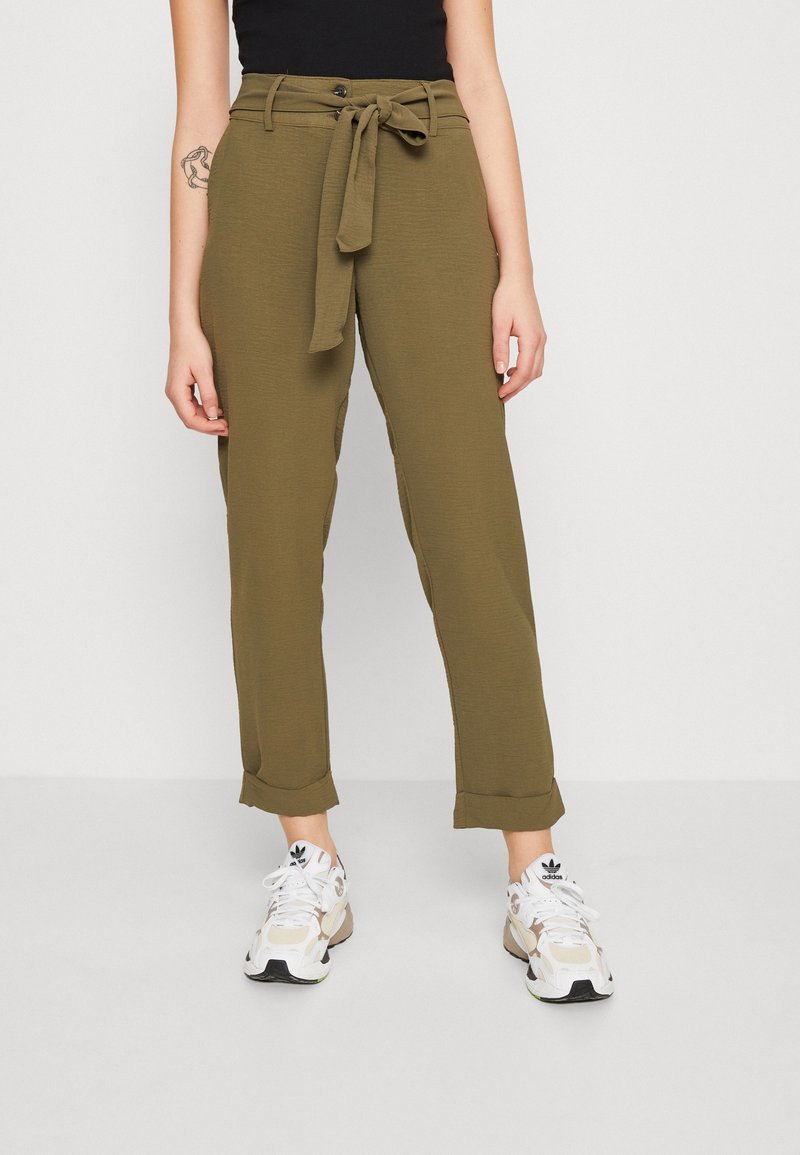Personne portant un pantalon paperbag taille haute vert olive avec une ceinture en tissu nouée devant, associé à des baskets blanches Adidas.