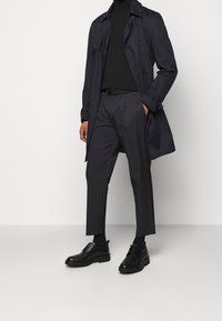 Cappotto trench blu navy su un dolcevita nero, abbinato a pantaloni scuri su misura e scarpe nere con i lacci, caratterizzato da un design elegante e da minimi accenti.