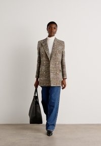 Doppelt knöpfbarer Tweed-Blazer in Brauntönen, Grün und Pink, kombiniert mit einem weißen Rollkragenpullover und blauen weiten Jeans; trägt eine schwarze Tote-Bag.