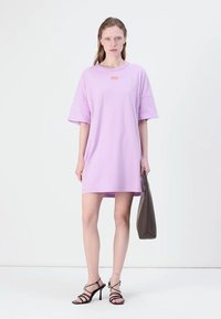 Lilla oversized t-skjortekjole med korte ermer, med en liten oransje logo. Modellen har på seg svarte strappy stilettos og bærer en grå veske.