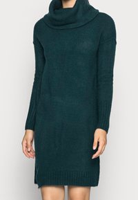 Abito in maglia teal con scollo a barchetta, maniche lunghe a costine e una finitura testurizzata. Presenta spacchi laterali all'orlo per garantire libertà di movimento.
