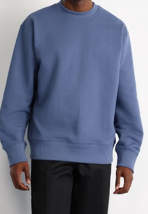 Mann trägt ein blaues Sweatshirt mit rundem Ausschnitt, gerippten Bündchen und Saum, kombiniert mit schwarzen Hosen vor hellem, einfarbigem Hintergrund.