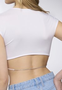 Top bianco a taglio crop con maniche corte, abbinato a jeans a vita alta di colore blu chiaro. Una catena di strass argentata accentua la vita.