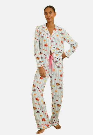 Žena oblečená v svetlomodrých pruhovaných pyžamách s farebnými ilustráciami jedál a ružovým šnúrkovým pásom, stojaca bosá na bielom pozadí.