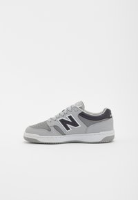 Šedé a černé nízké tenisky New Balance s tkaničkami, perforovanou špičkou a bílou podrážkou, zobrazené z boku na bílém pozadí.