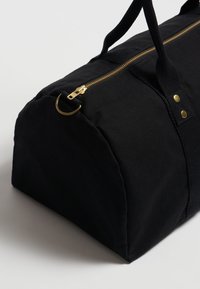 Černá plátěná taška přes rameno s zlatým zipem, kulatým designem, silnými popruhy a knoflíkovými akcenty. Hladký povrch s matným finišem.
