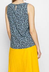 Haut sans manches bleu avec un imprimé floral noir. Présente une fermeture à l'arrière en forme de goutte. Associé à une jupe fluide jaune.