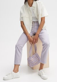 Chemise crème à manches courtes, débardeur blanc, pantalon taille haute lavande, baskets blanches et sac à main tissé violet sur un bloc de bois.