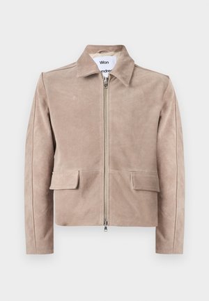 Mladić s kovrčavom kosom koji nosi bež jaknu na patent zatvarač, crnu košulju, široke plave traperice i crne cipele, stoji s jednom rukom u džepu.