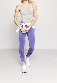 Hellgrauer Camouflage-Sport-BH mit 'PUMA'-Logo, kombiniert mit lila strukturierten Leggings mit Mesh-Elementen. Weiße Sneakers runden das Outfit ab.