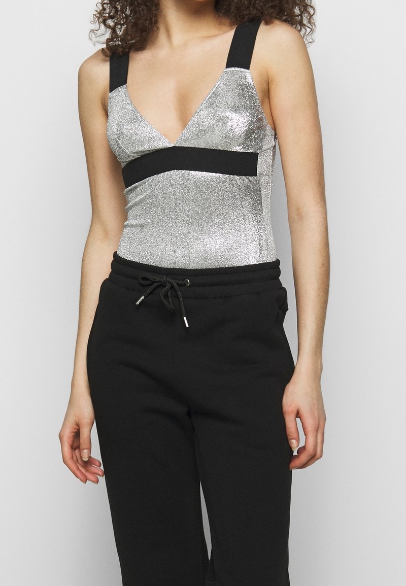 Femme portant un haut sans manches argenté pailleté avec des bretelles et une ceinture noires, associé à un pantalon noir à cordon de serrage.