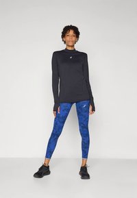 Woman standing in black long-sleeve top and blue patterned leggings with black athletic shoes against a plain light background.