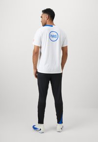 White short-sleeve shirt with blue accents, featuring the word "PARIS" in a circular design on the back. Worn with black pants and cleats.