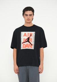 Μαύρο βαμβακερό T-shirt με μεγάλη γραφική απεικόνιση σε πορτοκαλί και λευκό που αναγράφει "AIR JORDAN" και έναν σιλουέτα τινόμενου προσώπου. Κοντά μανίκια.