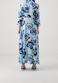Květinové maxi šaty v světle modré barvě s černými, modrými a zelenými vzory. Mají zapínání na knoflíky na zádech a tříčtvrteční rukávy, doplněné šedými podpatky.