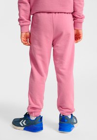 Pantalones deportivos rosas con textura suave, puños elásticos y un ajuste holgado, combinados con zapatillas azules y grises con detalles en blanco.