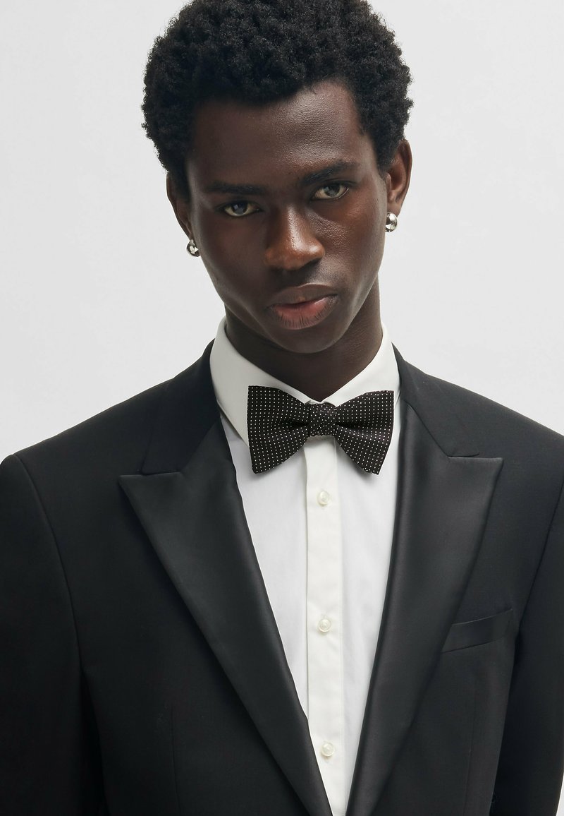 Zwarte tuxedo met satijnen revers, witte dress shirt en een zwart met witte stippen vlinderdas. Model heeft kort, krullend haar en een niet-reflecterende oorbel.