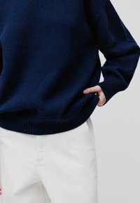 Tengerészkék kötött pulóver, bordás szegéllyel és mandzsettákkal, fehér nadrággal párosítva. Az anyag lágy és texturált.