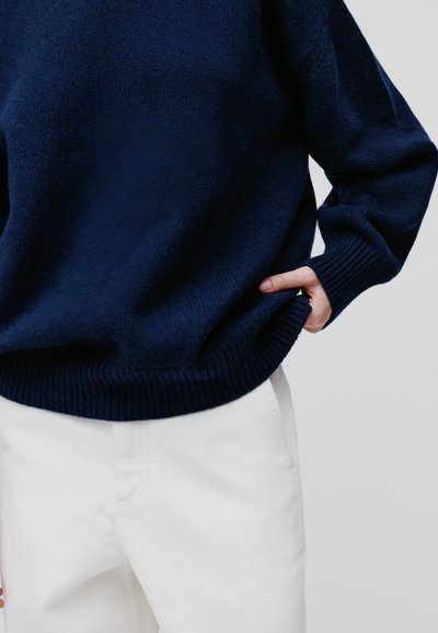 Navyblauwe gebreide trui met een geribde zoom en manchetten, gecombineerd met witte broek. De stof lijkt zacht en textuurrijk te zijn.