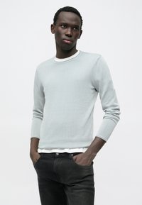 Camisola cinza clara com gola redonda e mangas compridas, combinada com jeans pretos. A camisola tem uma textura suave e um design ajustado.