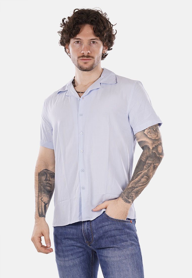Camicetta azzurra chiara a maniche corte con colletto a revers, realizzata in tessuto liscio, abbinata a jeans scuri. Presenta tatuaggi visibili.