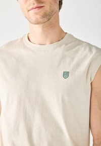 Light beige sleeveless t-shirt made of soft fabric, featuring a round neckline and a small green embroidered shield logo on the chest.