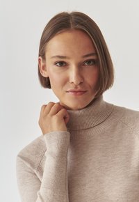Maglione beige a collo alto realizzato in morbido tessuto a costine, con maniche lunghe e una vestibilità aderente, che mostra un aspetto caldo e texturizzato.